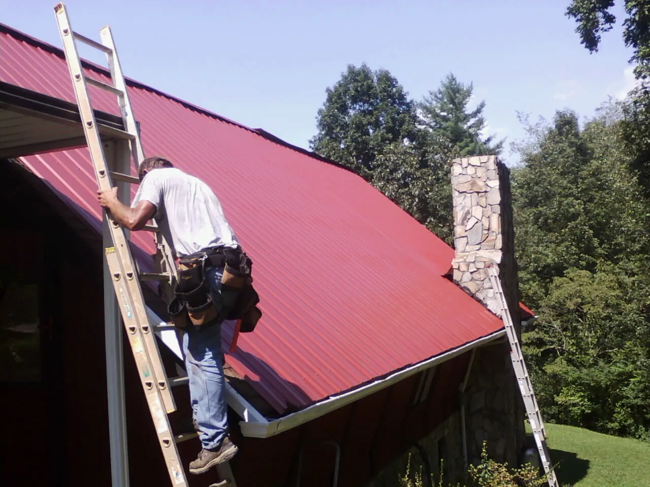 Roofer on a ladder beside a red metal roof and stone chimney — Soffit & Fascia Repair by Roofing Brothers in Rock Hill